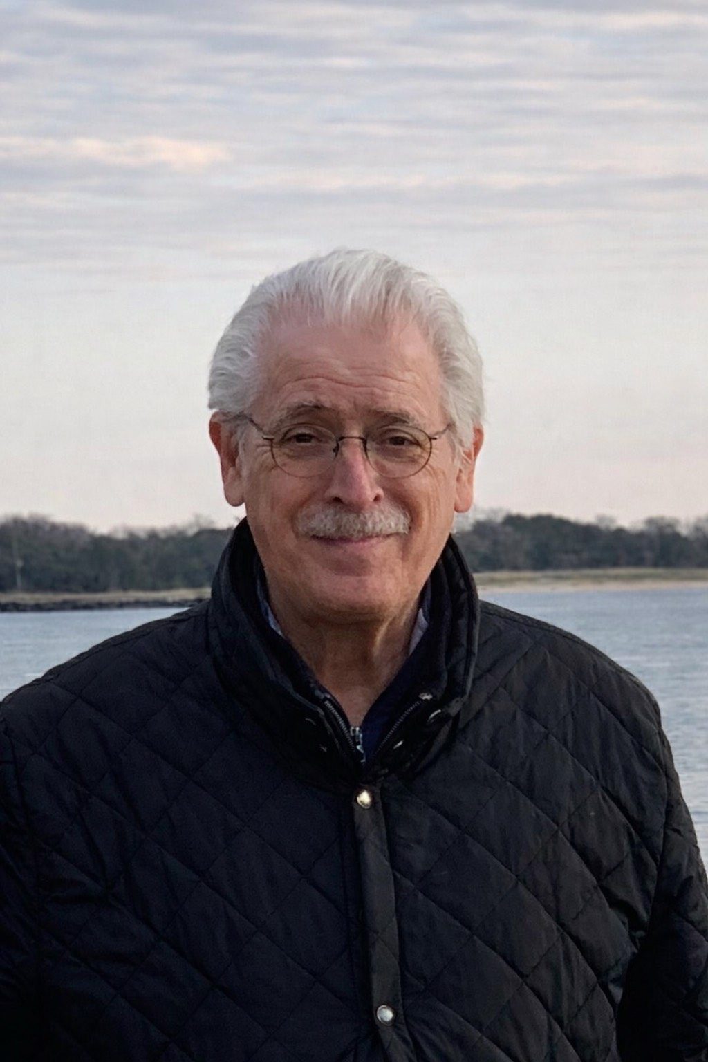 Elderly man with white hair wearing glasses and a dark jacket by a waterfront.